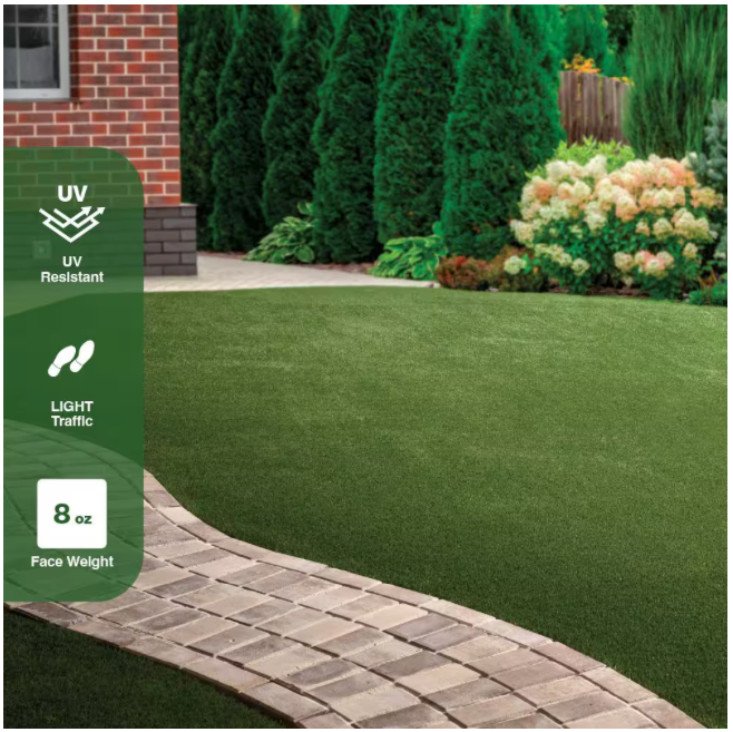 A landscaped yard features the TrafficMaster Emerald Green Precut Turf 6 ft. x 8 ft. Artificial Grass Rug, with a curved stone path and bushes by a brick house. Icons show UV resistance, light traffic suitability, and 8 oz face weight.