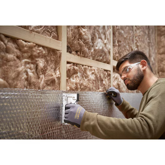 A man wearing safety glasses and gloves uses a STANLEY® Light-duty Steel Staple Gun to fasten reflective insulation to a wall with exposed fiberglass insulation and wooden framing.
