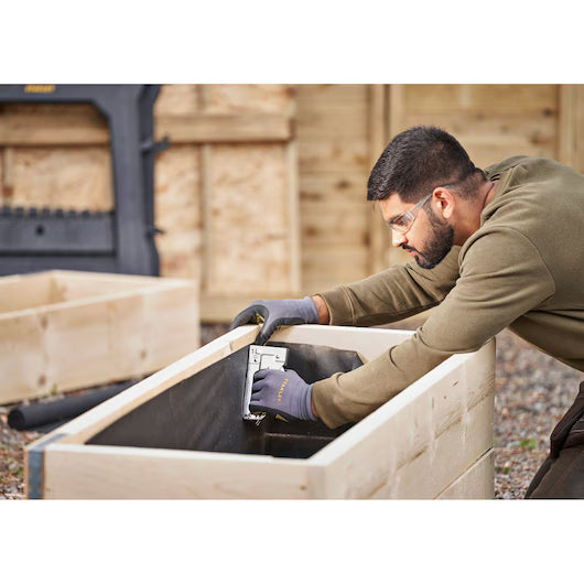 Wearing safety glasses and gloves, a man uses a STANLEY® Light-duty Steel Staple Gun by STANLEY to fasten black fabric to the interior of a wooden garden planter box outdoors.