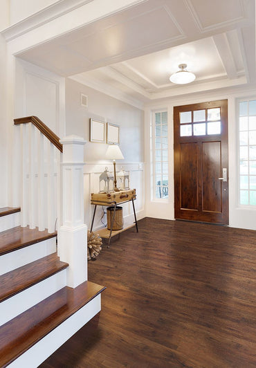A bright, modern entryway features MSIs Cyrus Braly Luxury Vinyl Planks (23.77 sq ft/box) in dark wood, a wooden front door with glass panels, white walls, staircase, console table with décor and lamp—all filled with natural light.