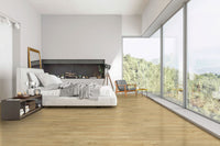 Modern bedroom with large bed, nightstand, and armchair boasts floor-to-ceiling windows overlooking forest. MSI Cyrus Brookline Luxury Vinyl Planks (23.77 sqft/box) offer 100% waterproof, click-lock installation for a minimalist look.