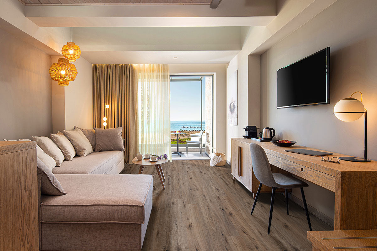 A modern hotel room with a sofa, desk, wall-mounted TV, and pendant lights. Sliding glass doors lead to a sea-view balcony. Warm neutral tones and MSI Cyrus Fauna Luxury Vinyl Planks create a cozy feel with durable CrystaLux™ protection.