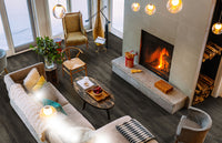 A cozy living room with a lit fireplace, modern furniture, beige sofa, Cyrus Billingham Luxury Vinyl Planks by MSI featuring waterproof SPC core and easy click-lock installation, wooden coffee table, large armchair, tall windows, and stacked logs nearby.