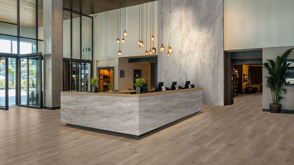 Modern hotel lobby with a large marble reception desk, pendant lights, tall windows, indoor plants, and matte beige wood-look MSI Cyrus Cranton Luxury Vinyl Planks (100% waterproof CrystaLux layer), creating a bright, open atmosphere.