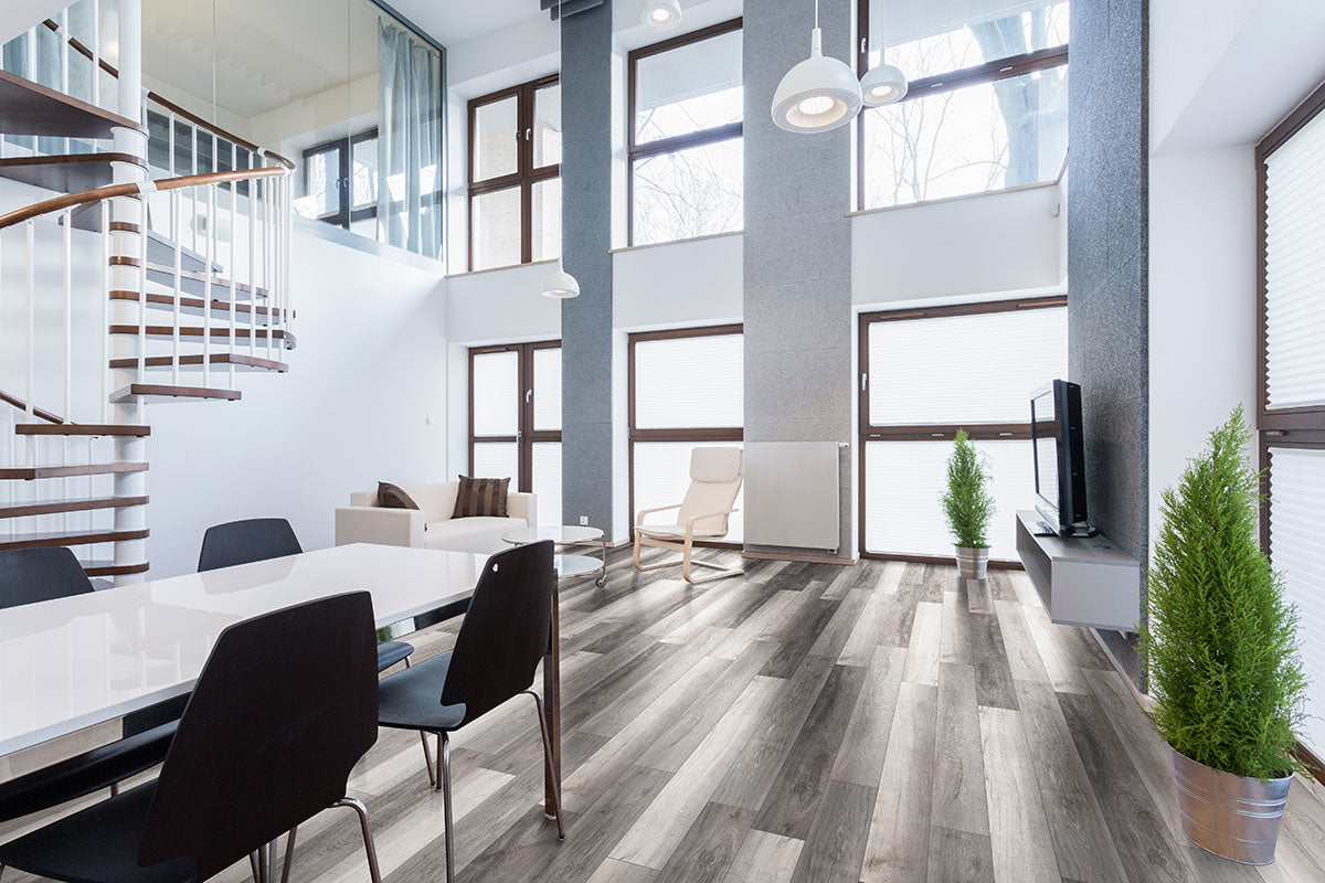 Modern living room with high ceilings, large windows, a spiral staircase, minimalist furniture, a dining table with black chairs, TV, two potted plants, and MSI Cyrus Draven Luxury Vinyl Planks featuring easy click-lock installation.