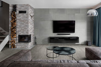 Modern living room with a gray sectional, wall-mounted TV, black shelves, round coffee tables, fireplace with stacked firewood, large windows with blue curtains, and durable MSI Cyrus Grayton Luxury Vinyl Planks flooring for waterproof protection.