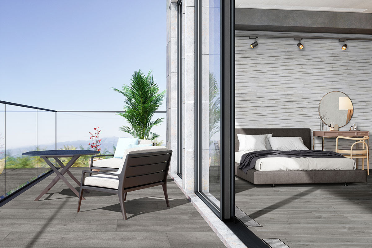 A modern bedroom with MSI Katella Luxury Vinyl Planks flooring opens to a balcony with glass railing, two chairs, and a round table. Inside are a gray bed, wooden furniture, a decorative wall, and a large potted plant outside.
