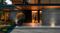 A person approaches a modern homes entrance at dusk, lit by warm lights. The house showcases stone, wood, large windows, and MSI Katella Luxury Vinyl Planks with click-lock waterproof flooring near the landscaped walkway.