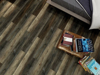 A bedside table with stacked books and an armchair sits next to a bed with gray bedding on an MSI Cyrus Stable Luxury Vinyl Planks floor, showcasing an antique hardwood look in dark and light brown tones.
