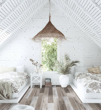 A bright attic bedroom with two beds, featuring MSI Cyrus Weathered Brina Luxury Vinyl Planks for a rustic wood look, white wooden walls and ceiling, large window with greenery, patterned bedding, woven pendant light, and potted plants.