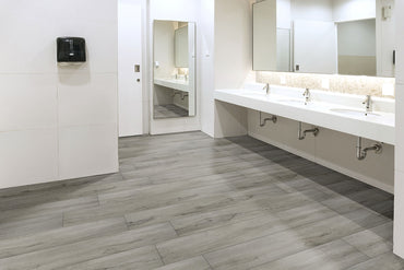 A clean, modern public restroom with MSI XL Cyrus Dunite Oak Luxury Vinyl Planks flooring (22.44 sq ft/box), featuring a waterproof SPC core and CrystaLux™ wear layer, three sinks with mirrors, white tiled walls, and a black towel dispenser.