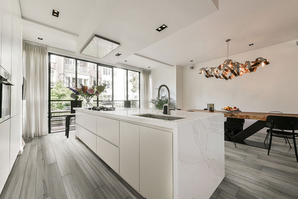 Modern kitchen with a large white marble island, MSI XL Cyrus Dunite Oak Luxury Vinyl Planks floors featuring a CrystaLux™ wear layer, large windows with sheer curtains, and a contemporary dining area with a sculptural light fixture.