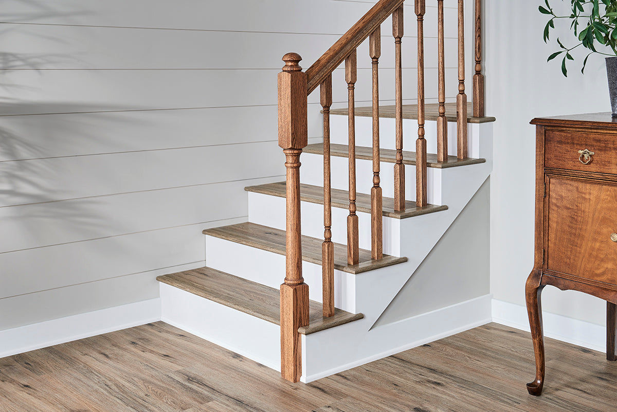 A wooden staircase with brown steps, white risers, and a wooden handrail stands beside a chest of drawers on MSI’s Cyrus Fauna Luxury Vinyl Planks flooring (23.77 SQFT box), featuring CrystaLux™ wear-layer protection. A plant appears in the top right.