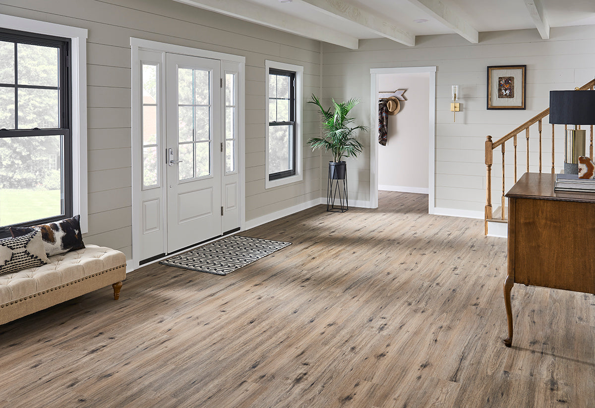 Bright entryway with MSI Cyrus Fauna Luxury Vinyl Planks flooring (23.77 SQFT/box) featuring CrystaLux™ wear-layer protection, white glass-panel front door, black-framed windows, striped rug, beige bench, potted plant, and wooden stair railing.