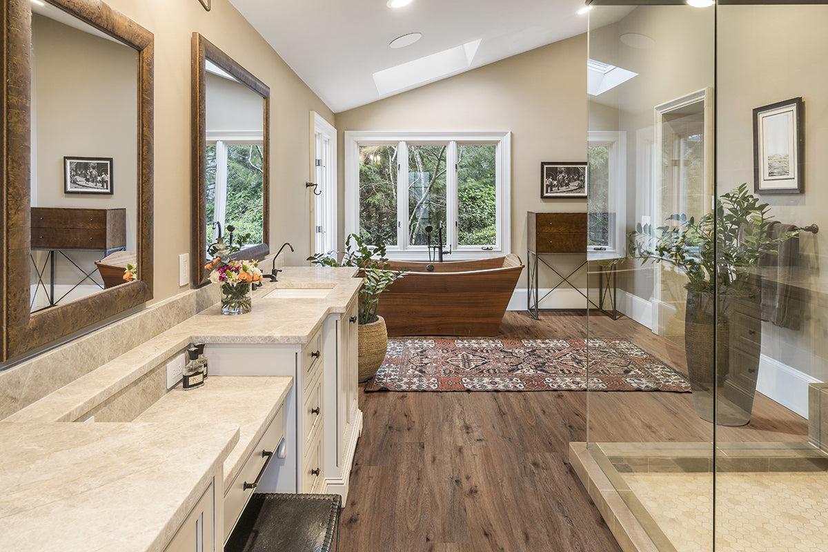 Spacious bathroom with double sinks, large mirrors, a freestanding wooden tub, glass shower, indoor plants, and a colorful rug on MSI Cyrus Fauna Luxury Vinyl Planks (BOX 23.77 SQFT) flooring featuring CrystaLux™ wear-layer protection. Large windows provide natural light.
