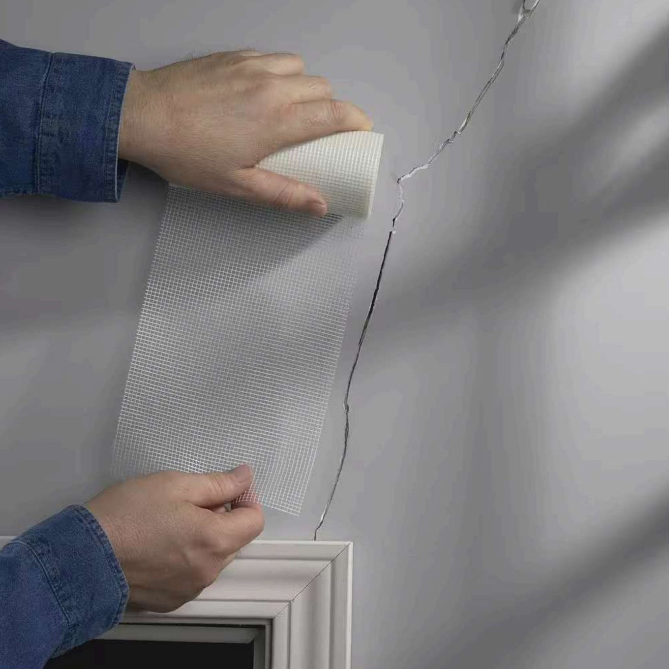 A person applies ADFORS FibaTape FDW6568-U 6 in. x 75 ft. Crackstop Self-Adhesive Wide Wall Repair Tape over a crack in a gray wall above a window frame, preparing the area for further repair work.