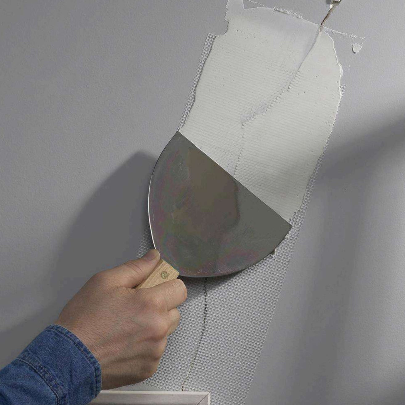 A hand uses the ADFORS FibaTape FDW6568-U 6 in. x 75 ft. Crackstop Self-Adhesive Wide Wall Repair Tape and joint compound to smooth a drywall seam on a gray wall during repair.