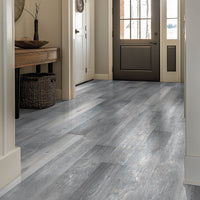 A hallway features MSIs Cyrus Finely Luxury Vinyl Planks (100% waterproof, gray and greige wood tones) flooring, a dark glass-panel front door, a console table with decor, and a woven basket below. Natural light streams in.