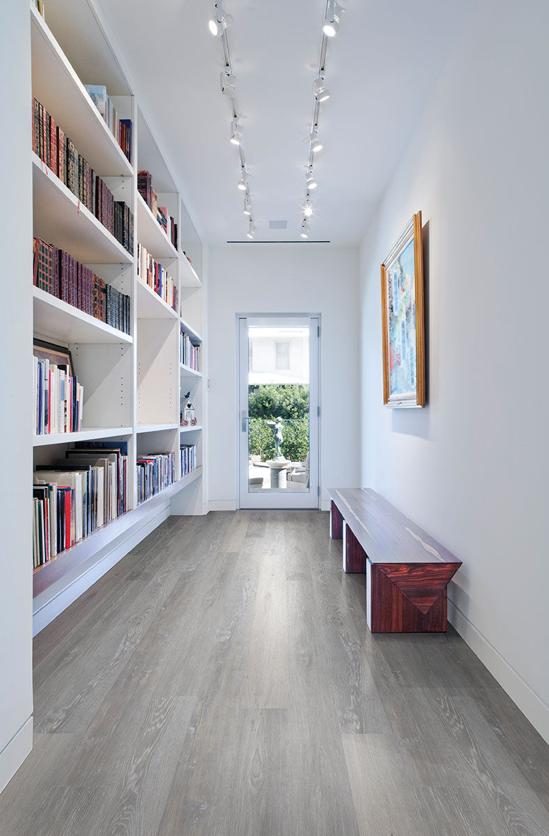 A modern hallway featuring MSI Cyrus Finely Luxury Vinyl Planks, built-in bookshelves on the left, a wooden bench on the right in gray and greige tones, track lighting overhead, and a glass door opening to lush greenery outside.