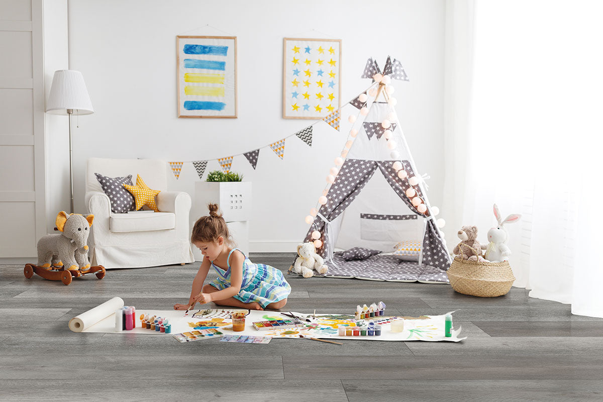 A young girl paints with watercolors in a bright playroom, sunlight illuminating MSI XL Cyrus Katella Ash Luxury Vinyl Planks flooring. Plush toys and colorful wall art add charm near her tent draped with string lights.