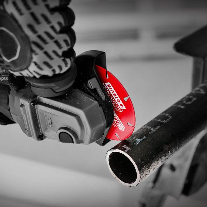 A person wearing patterned gloves uses the Diablo DIABLO DDD070DIA101F 7 in. Diamond Metal Cut-Off Disc to cut a black metal pipe with a circular saw. The durable red blade, marked with white text, stands out against the blurred grayscale background.