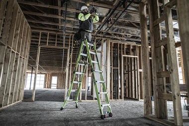 A construction worker in safety gear stands on a LITTLE GIANT 17117 CONQUEST 2.0 1A 7.5-15 F/G ARTICULATED EXTENSION LADDER, working on exposed wiring inside a building with unfinished wooden framing.