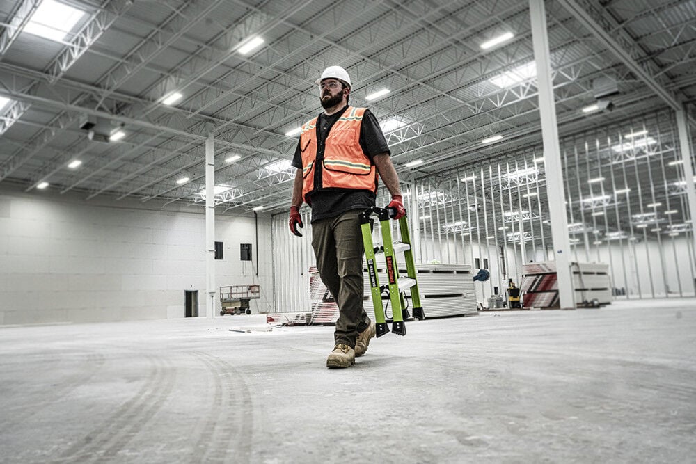 A construction worker in a white hard hat and orange vest carries a LITTLE GIANT 11953 Sure Step 1AA 3-Step Double-Sided Fiberglass Step Stool across a large indoor site with high ceilings, exposed beams, and unfinished walls.