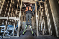 A construction worker in an orange safety vest stands on a LITTLE GIANT 11953 Sure Step 1AA 3-Step Double-Sided Fiberglass Step Stool, working on the framing of a building with exposed wooden beams and unfinished walls.