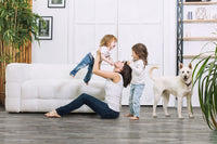 A woman sits on the floor holding a laughing toddler above her while another child and a white dog stand nearby in a bright living room with lush plants, a white sofa, and stylish MSI Cyrus Mezcla Luxury Vinyl Planks flooring.