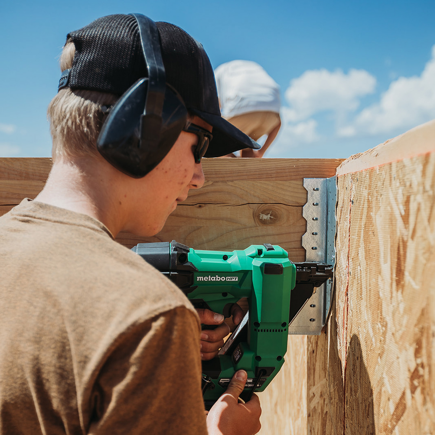 A person with ear protection and a cap uses the Go Build Metabo HPT NR1838DKA 18V MultiVolt Brushless Metal Connector Nailer to secure a metal bracket to wood outdoors. Another capped person is partially seen in the background.