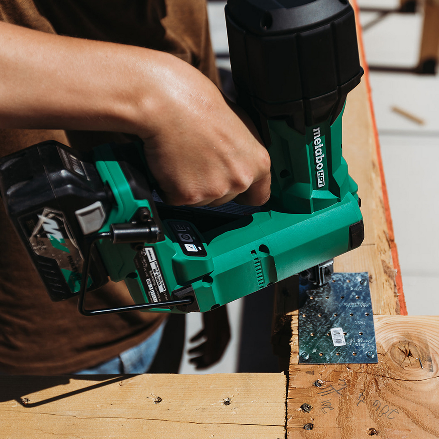 A person uses the Go Build Metabo HPT NR1838DKA 18V MultiVolt Brushless Metal Connector Nailer (1x4.0Ah) to attach a metal bracket to a wooden beam during construction.
