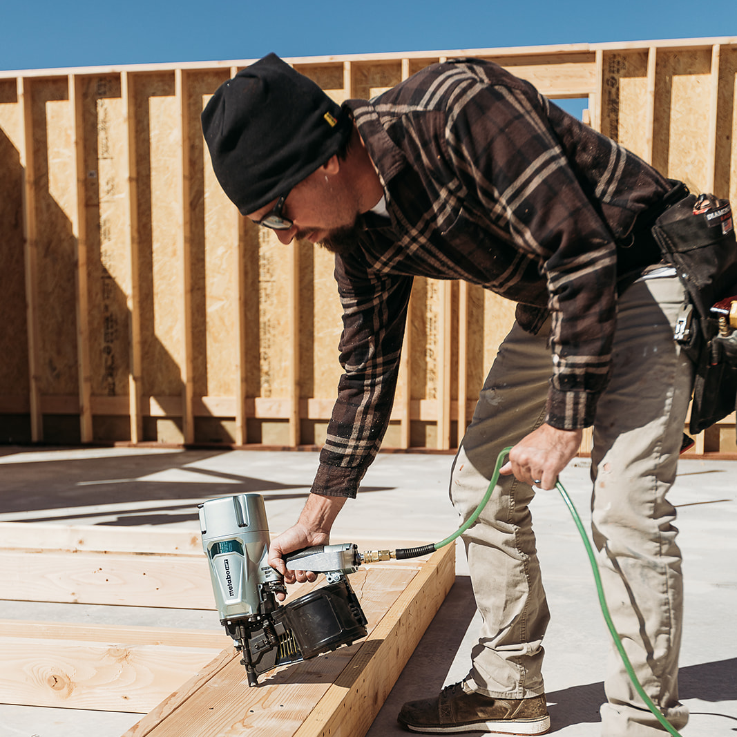 A construction worker in a beanie, sunglasses, and plaid shirt uses the Go Build Metabo HPT NV83A5 3-1/4 Inch Coil Framing Nailer to fasten wooden boards at a site with unfinished wood walls in the background.