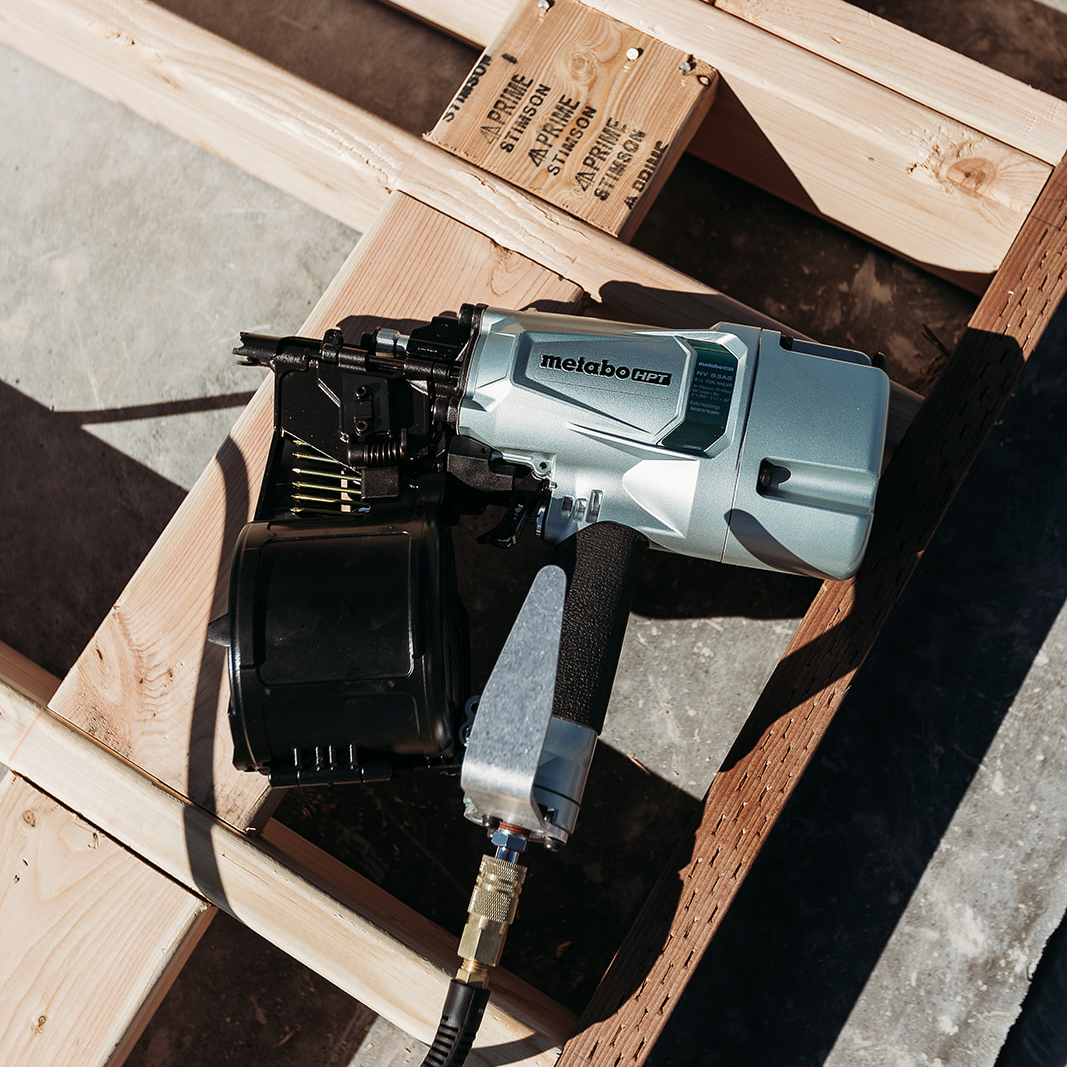 A close-up of a Metabo HPT NV83A5 | 3-1/4 Inch Coil Framing Nailer by Go Build, The Fastest Way To Build, rests on a partially assembled wooden frame with sunlight casting shadows over the wood and concrete below.