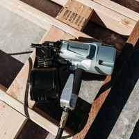 A close-up of a Metabo HPT NV83A5 | 3-1/4 Inch Coil Framing Nailer by Go Build, The Fastest Way To Build, rests on a partially assembled wooden frame with sunlight casting shadows over the wood and concrete below.