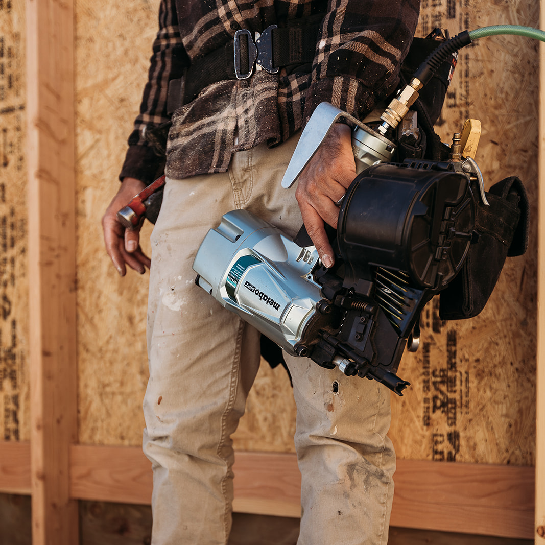 A construction worker in khaki pants and a plaid shirt wields the Metabo HPT NV83A5 | 3-1/4 Inch Coil Framing Nailer by Go Build, The Fastest Way To Build, tool belt on waist and air hose attached, ready for precision beside a wooden frame wall.