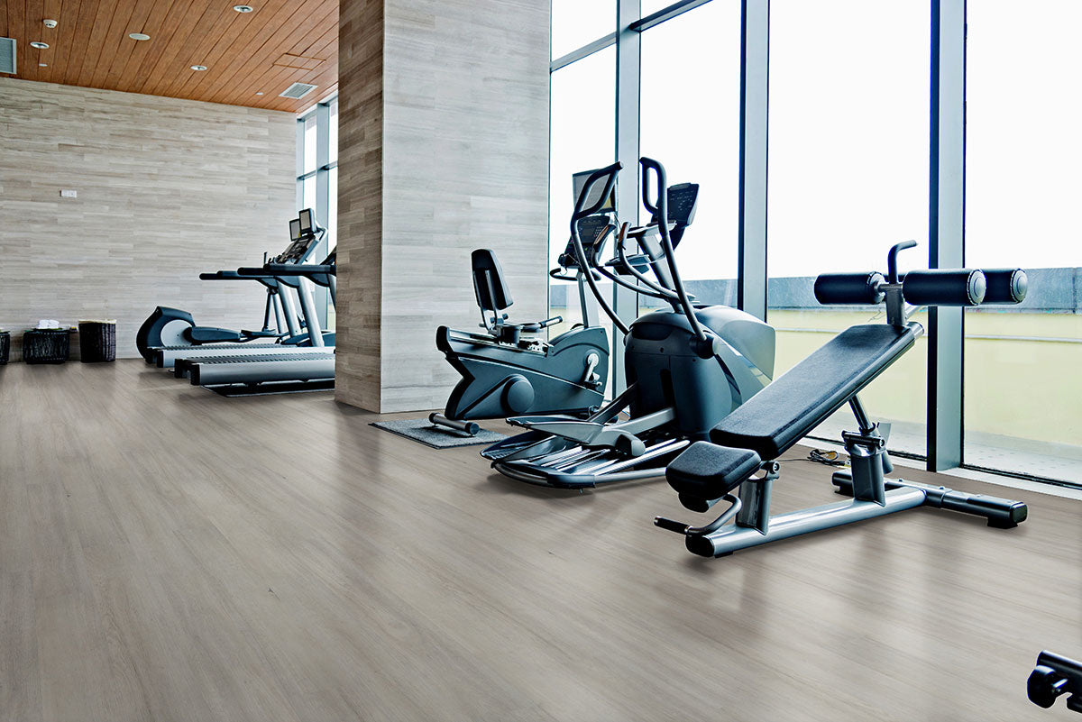 A modern gym with large windows features treadmills, an elliptical, a stationary bike, and a weight bench atop MSIs Cyrus Brianka Luxury Vinyl Planks, filling the bright space with style and natural light.
