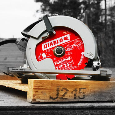A circular saw featuring the DIABLO D0724A 7-1/4 in. x 24-Teeth Framing Saw Blade for Wood, renowned for its Extreme Cutting Efficiency, rests on a wooden plank. The predominantly grayscale backdrop accentuates the vibrant red and silver of this Diablo Framing Saw Blade. Trees and various construction materials surround the scene.
