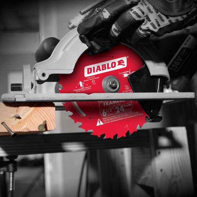 A close-up of a Diablo circular saw fitted with the DIABLO D0624A 6-1/2 in. x 24-Teeth Framing Saw Blade effortlessly slices through wood, highlighted by its vibrant red against the stark black-and-white background. The operator, with gloves on, expertly guides the tool atop a workbench.