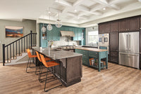 Modern kitchen with teal cabinets, dark wood island, stainless steel appliances, and three orange bar stools; MSI Cyrus Ryder Luxury Vinyl Planks flooring (SPC core) adds reclaimed wood warmth, with a staircase and artwork on the left.