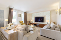 A spacious, modern living room with beige sofas, a glass coffee table, a green armchair, floor lamps, MSI Cyrus Sandino Luxury Vinyl Planks flooring (waterproof SPC core), a wall-mounted TV, and windows with curtains letting in natural light.