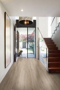 Modern entryway with MSI Cyrus Sandino Luxury Vinyl Planks (23.77 sqft/box), featuring a waterproof SPC core and CrystaLux™ protection, glass staircase, round hanging light, wall art, and a dog outside the glass front door with garden views.