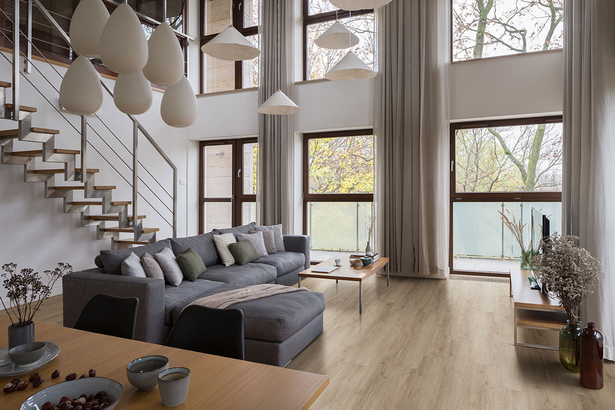 A modern living room features tall windows, a gray sectional, tables, TV, and pendant lights. MSI Cyrus Sandino Luxury Vinyl Planks BOX (23.77 SQFT) add an airy feel with light wood tones and waterproof SPC core under a metal-railed staircase.