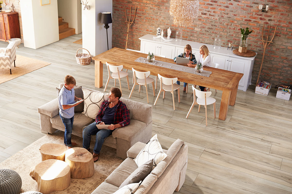 A cozy living and dining room features MSI’s XL Cyrus Akadia Luxury Vinyl Planks (22.44 SQFT) with neutral beige tones, brick walls, and warm decor. Two people chat on a couch while two others work at a long dining table.