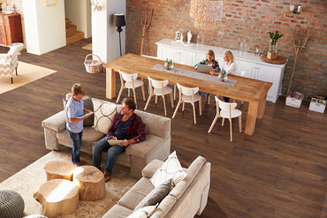 A spacious, modern living and dining room with stylish decor features MSI XL Cyrus Braly Luxury Vinyl Planks—100% waterproof flooring. Two men use tablets on the sofa while two girls work at a wooden dining table in the bright space.