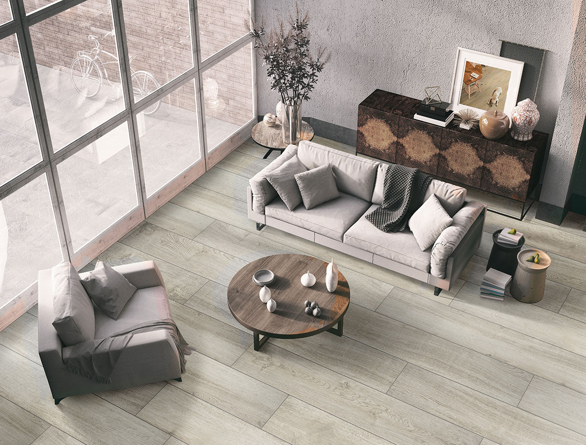 A modern living room features MSI’s XL Cyrus Brianka Luxury Vinyl Planks for durable, waterproof flooring. Large windows, a gray sofa set, wooden coffee table, dark sideboard with decor, and neutral tones create a cozy, airy atmosphere.
