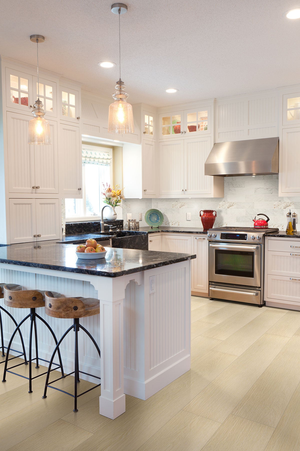 Bright kitchen with white cabinets, stainless steel appliances, a large black island with wooden bar stools, pendant lights, and decor—finished with MSI XL Cyrus Lenexa Creek Luxury Vinyl Planks featuring the durable CrystaLux™ wear layer.