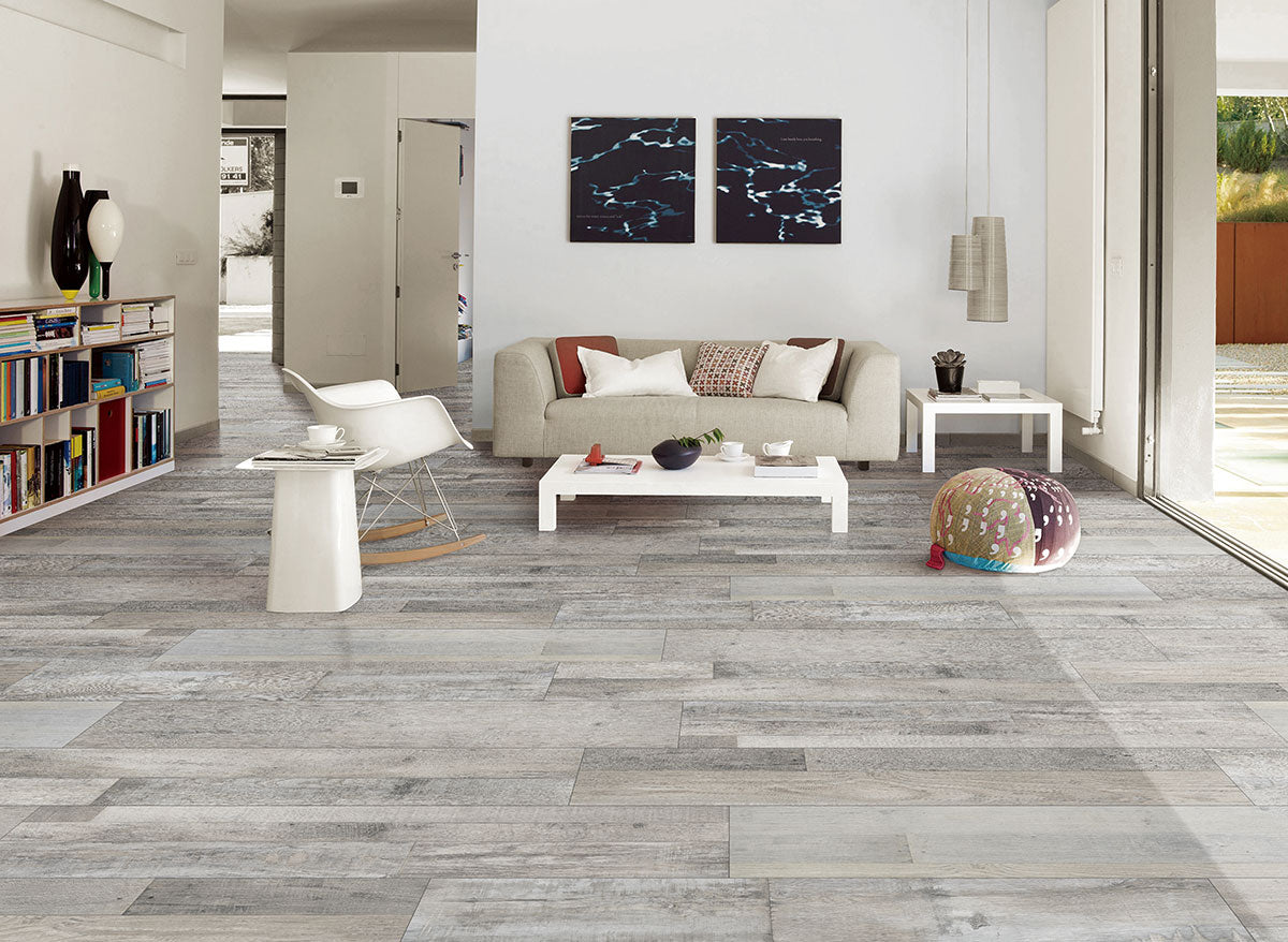 A modern living room featuring MSI XL Cyrus Mezcla Luxury Vinyl Planks flooring, a beige sofa, two abstract paintings on a white wall, bookshelves, a low coffee table, a white chair, and a colorful pouf near large windows.