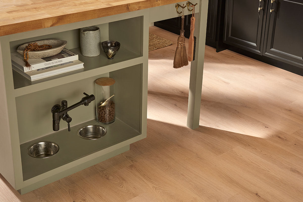 A kitchen island with a built-in pet feeding station, faucet, stainless steel bowls, kibble jar, and shelves for books and decor sits atop MSI XL Cyrus Runmill Isle Luxury Vinyl Planks flooring, complemented by sleek black cabinets.