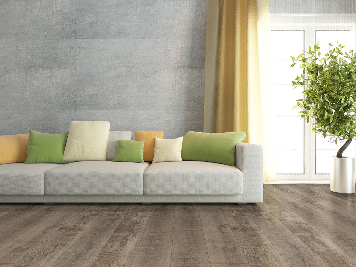 A modern living room features a light gray sofa, colorful pillows, and a textured wall. MSI XL Cyrus Ryder Luxury Vinyl Planks (22.44 SQFT/box) enhance the space, complemented by sheer yellow curtains and a fresh green plant.