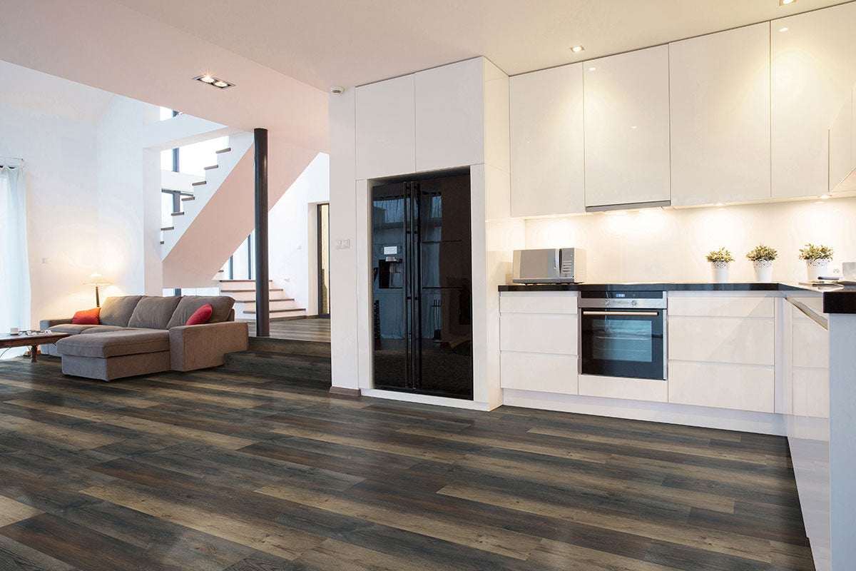 Modern open-plan kitchen and living area featuring white cabinets, black appliances, and MSI XL Cyrus Stable Luxury Vinyl Planks flooring with a durable CrystaLux wear layer. A gray sectional sofa anchors the space; stairs are in the background.